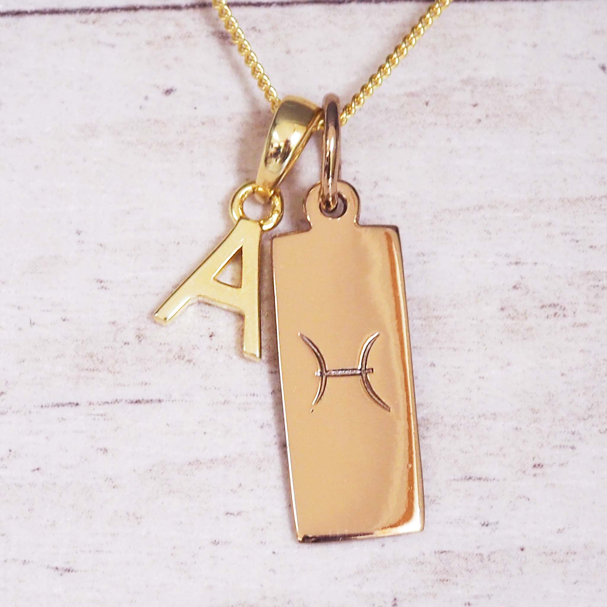 Close-up of gold necklace with A initial charm and rectangular tag engraved with the Pisces zodiac glyph on a fine chain