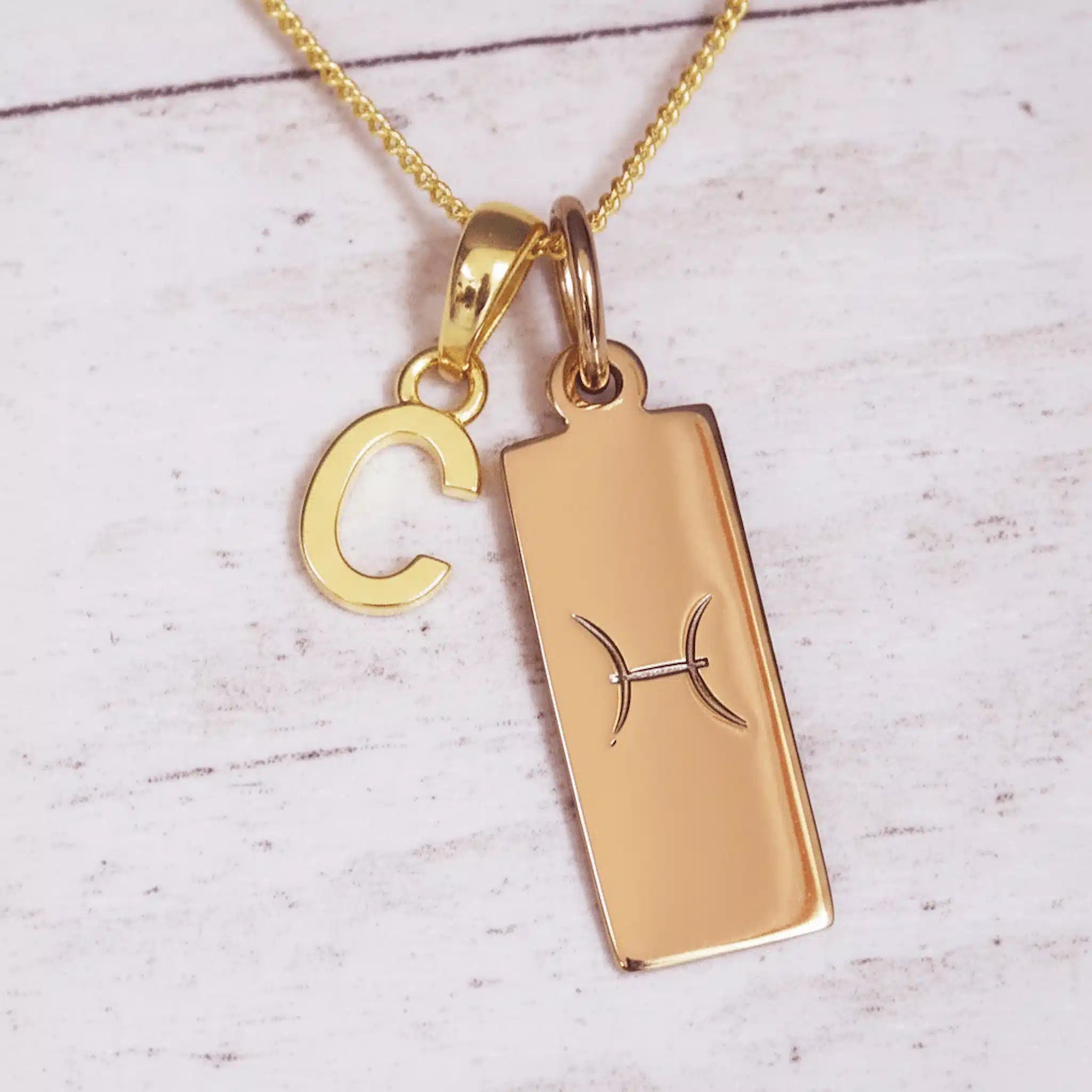 Gold necklace with a rectangular pendant and 'C' charm on a light wooden surface