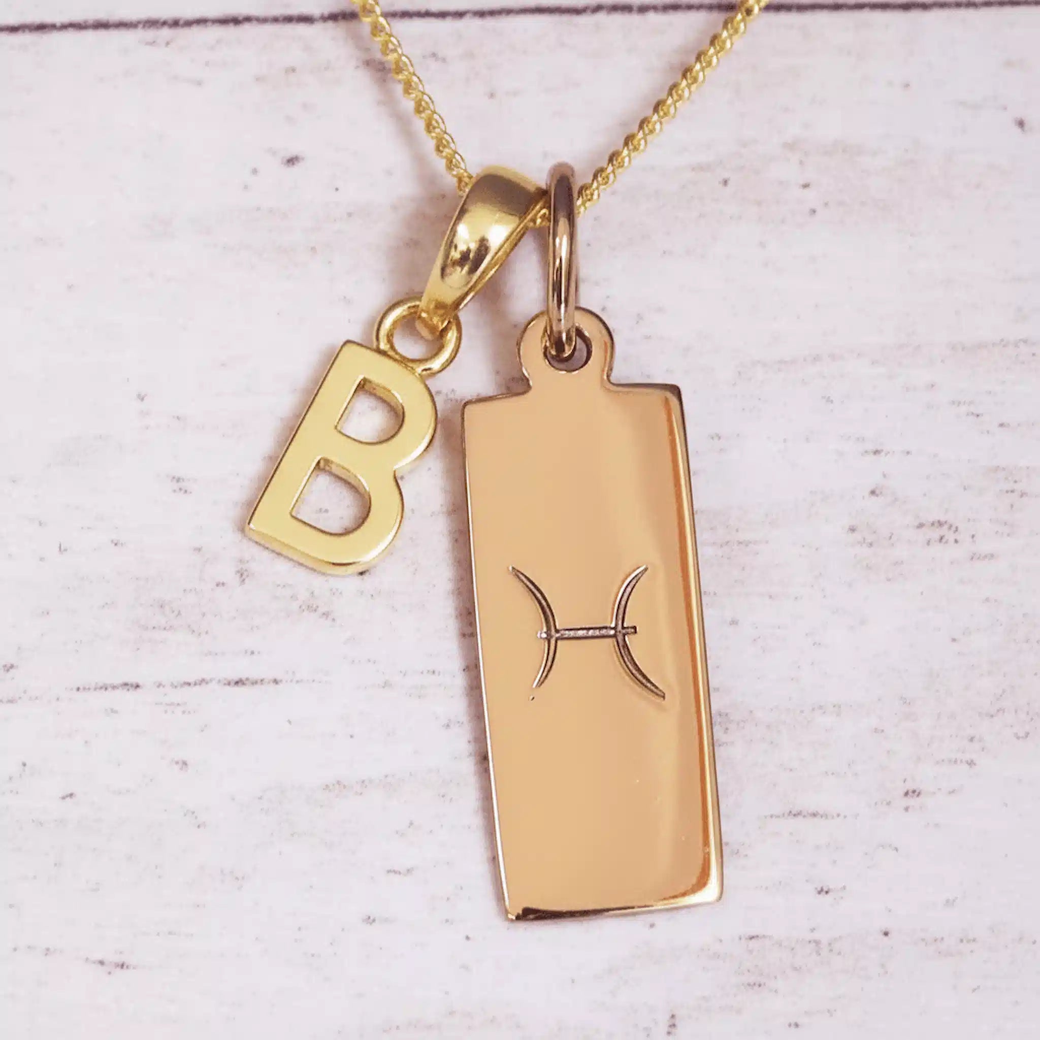 Gold necklace with 'B' pendant and zodiac sign on a light wooden background