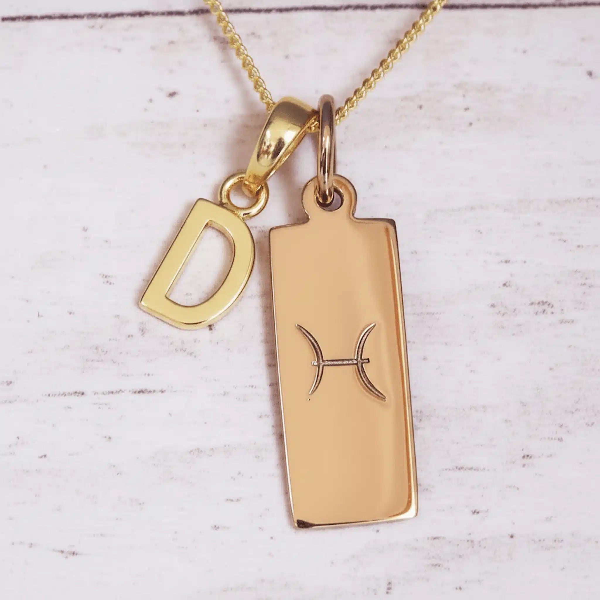 Gold necklace with a pendant featuring the letter 'D' and a zodiac symbol on a light wooden background.