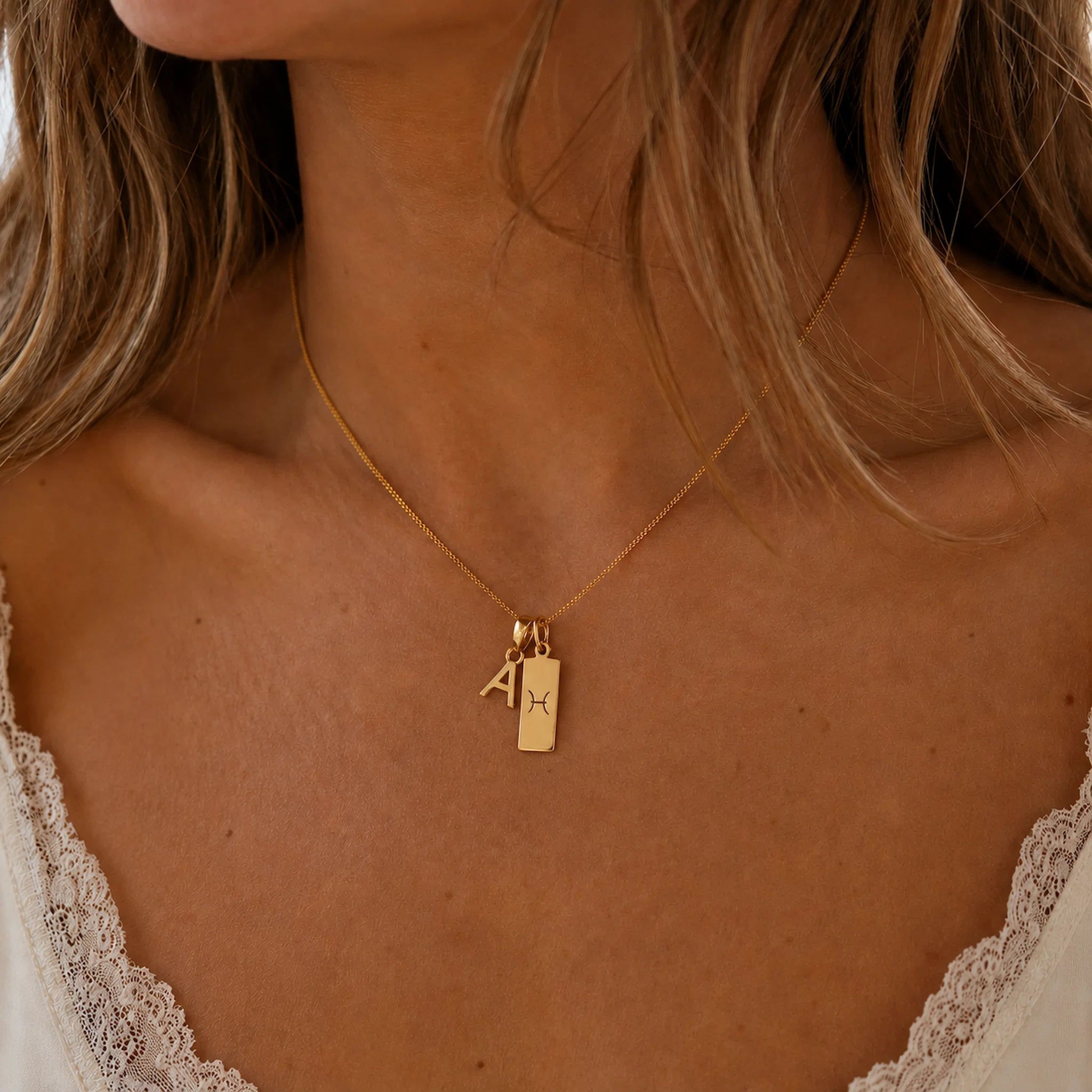 model wearing a gold necklace with a uppercase block letter pendant and a rectangle pendant with a pisces zodiac glyph.