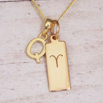 Gold initial Q and Aries zodiac bar necklace, close-up of the letter charm and engraved pendant on whitewashed wood