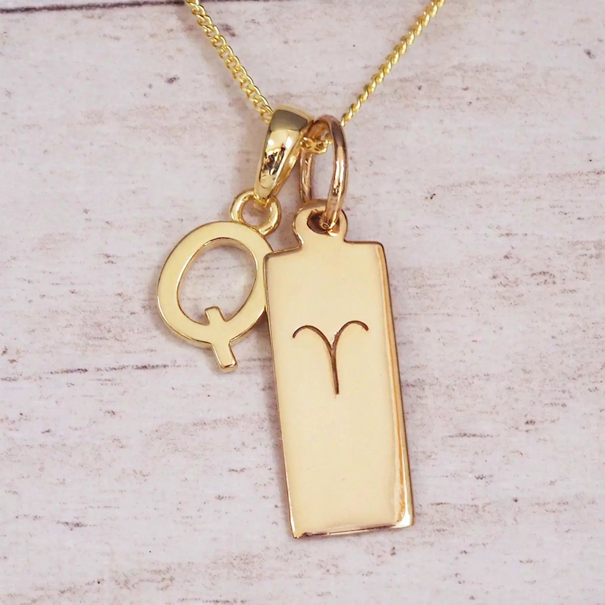 Gold initial Q and Aries zodiac bar necklace, close-up of the letter charm and engraved pendant on whitewashed wood