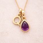 Gold Initial Q necklace with a purple teardrop amethyst pendant on a beige background