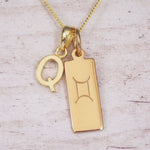 Gold initial Q and Gemini zodiac bar necklace, close-up of the letter charm and engraved pendant on whitewashed wood