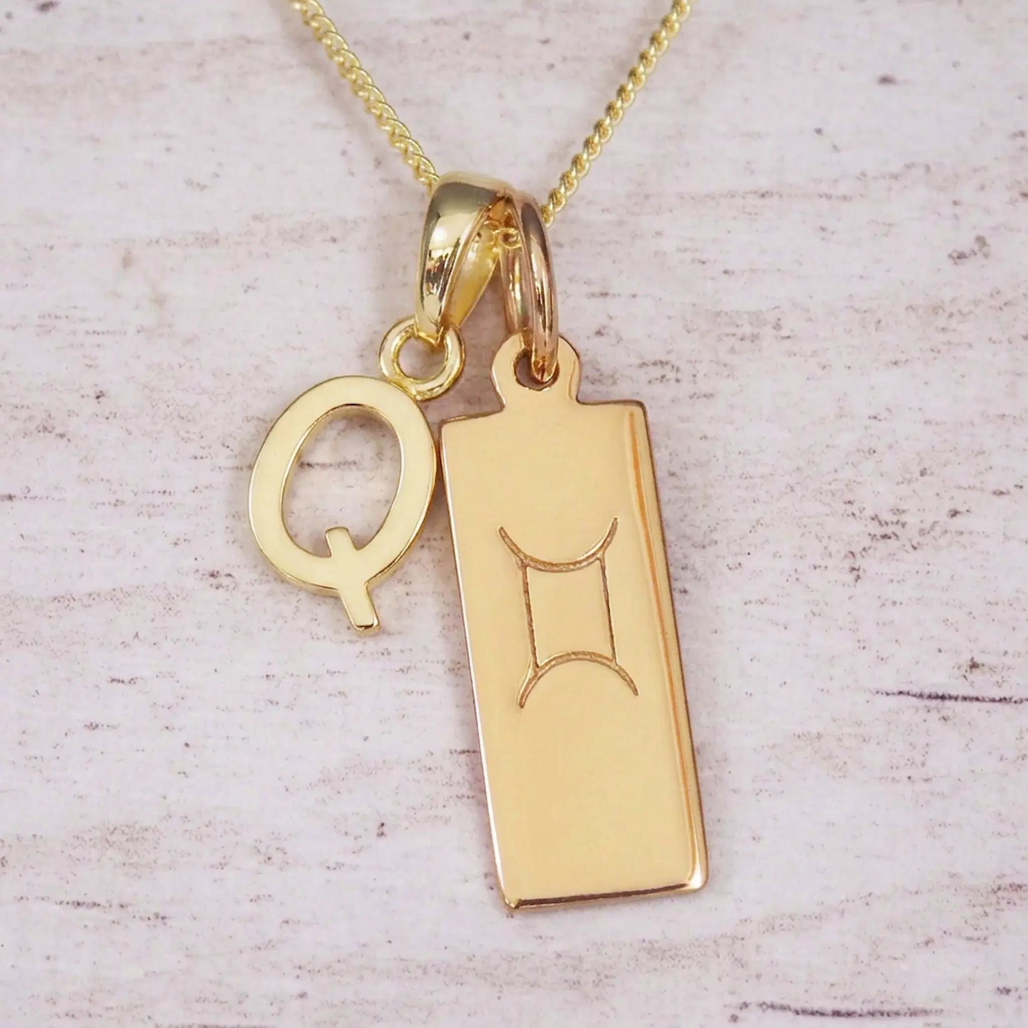 Gold initial Q and Gemini zodiac bar necklace, close-up of the letter charm and engraved pendant on whitewashed wood