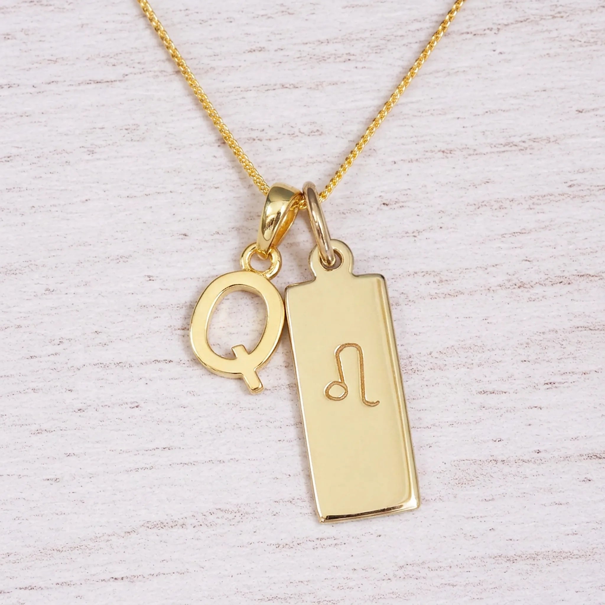 Gold initial and Leo zodiac necklace with a letter Q pendant and engraved Leo bar on white wood, close-up