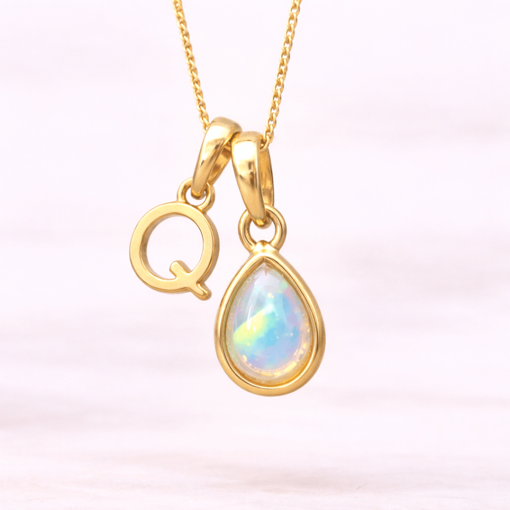 Gold necklace with 'Q' pendant and teardrop-shaped opal gemstone on a light pink background