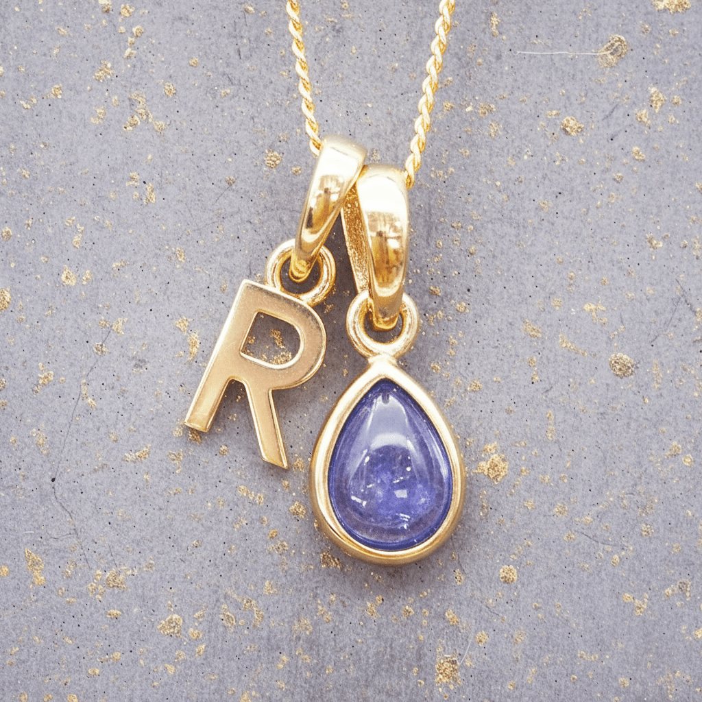 Gold necklace with 'R' pendant and teardrop-shaped purple tanzanite gemstone on a textured gray background.