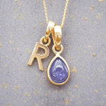 Gold December birthstone necklace with 'R' initial pendant and a natural tanzanite gemstone pendant on a grey background.