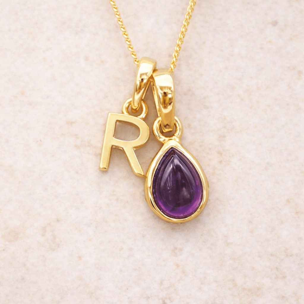 Gold necklace with 'R' pendant and purple teardrop amethyst charm on a beige background