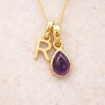 Gold necklace with 'R' pendant and purple teardrop amethyst charm on a beige background