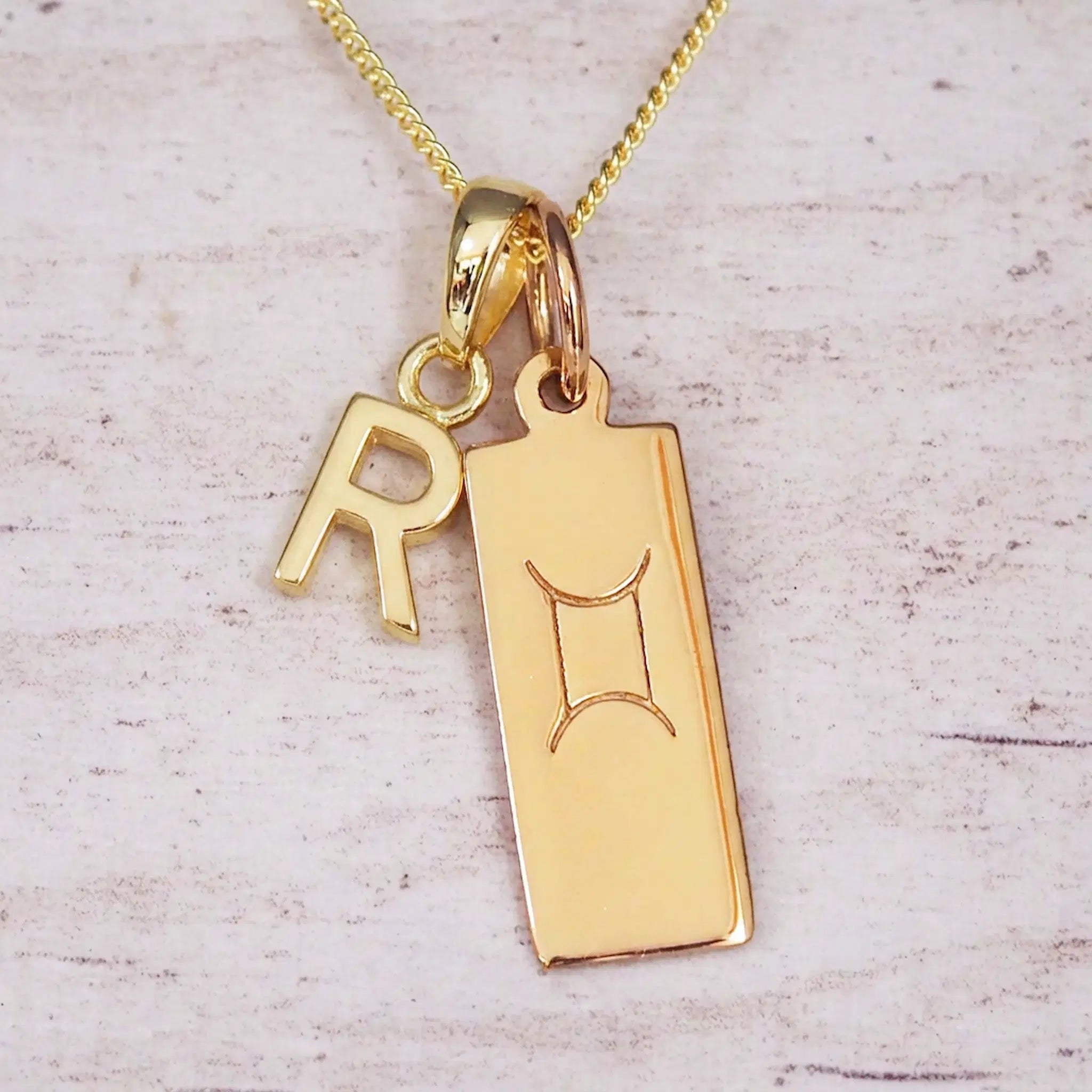 Gold initial R and Gemini zodiac bar necklace, close-up of the letter charm and engraved pendant on whitewashed wood