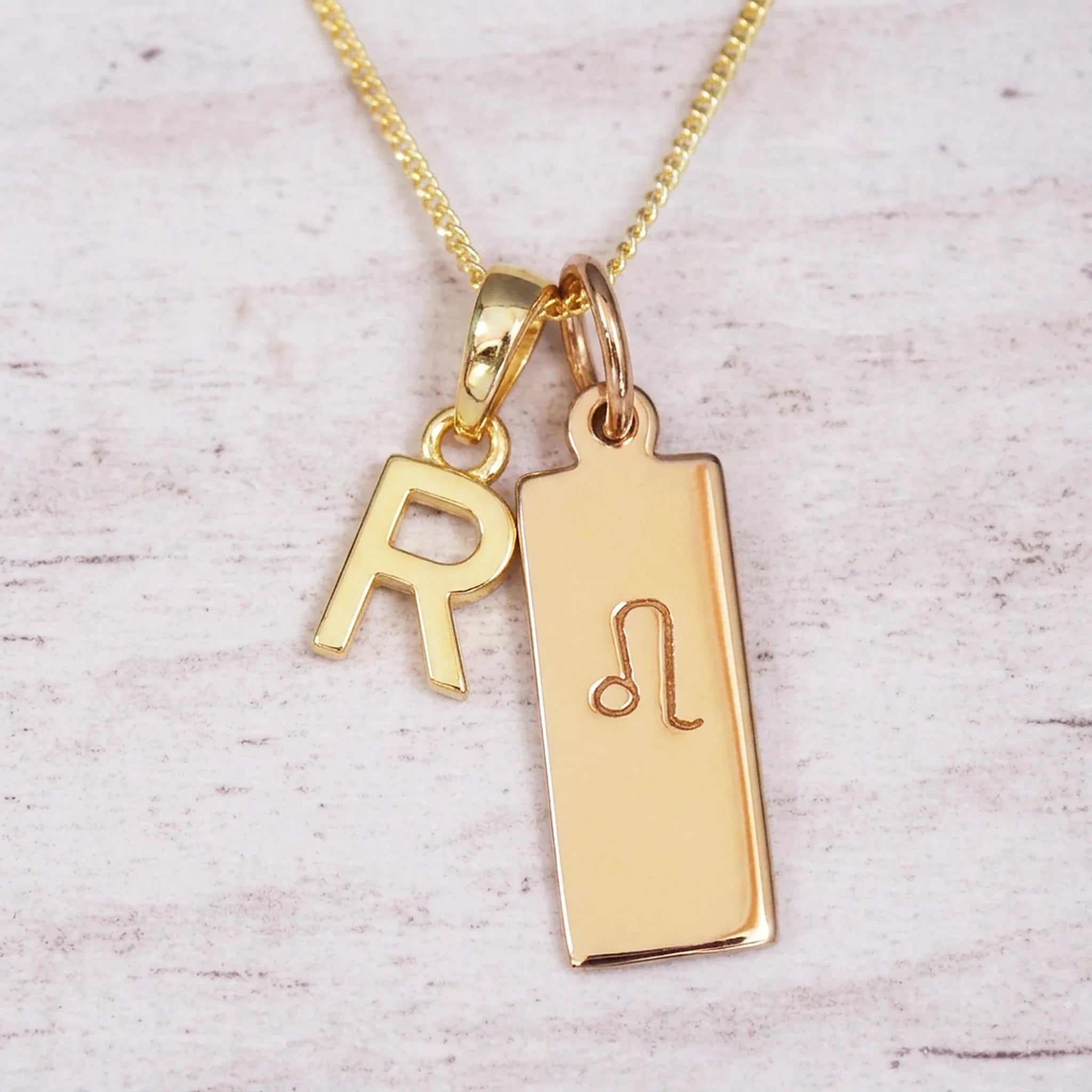 Gold initial and Leo zodiac necklace with a letter R pendant and engraved Leo bar on white wood, close-up