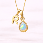 Gold necklace with 'R' pendant and teardrop-shaped opal on a light pink background