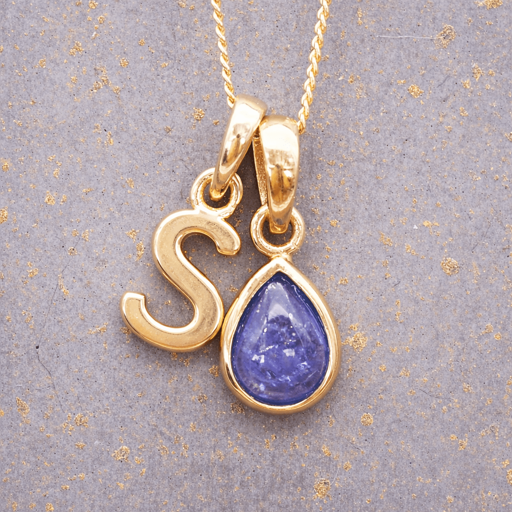 Gold necklace with 'S' pendant featuring a purple teardrop tanzanite gemstone on a textured gray background.