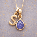 Gold necklace with 'S' pendant featuring a purple teardrop tanzanite gemstone on a textured gray background.