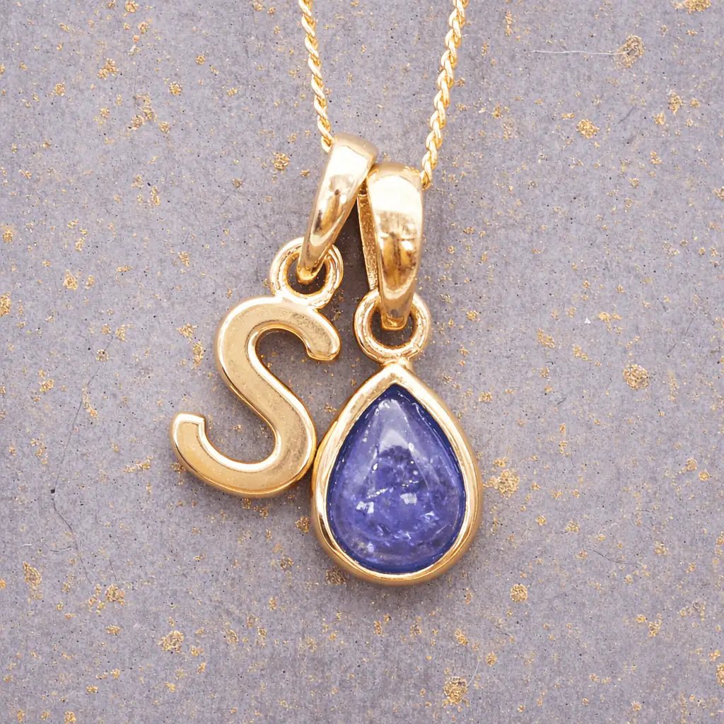 Gold December birthstone necklace with a pear-shaped natural tanzanite pendant and letter 'S' charm with a grey background.