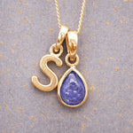 Gold December birthstone necklace with a pear-shaped natural tanzanite pendant and letter 'S' charm with a grey background.
