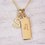 Gold initial and Leo zodiac necklace with a letter S pendant and engraved Leo bar on white wood, close-up