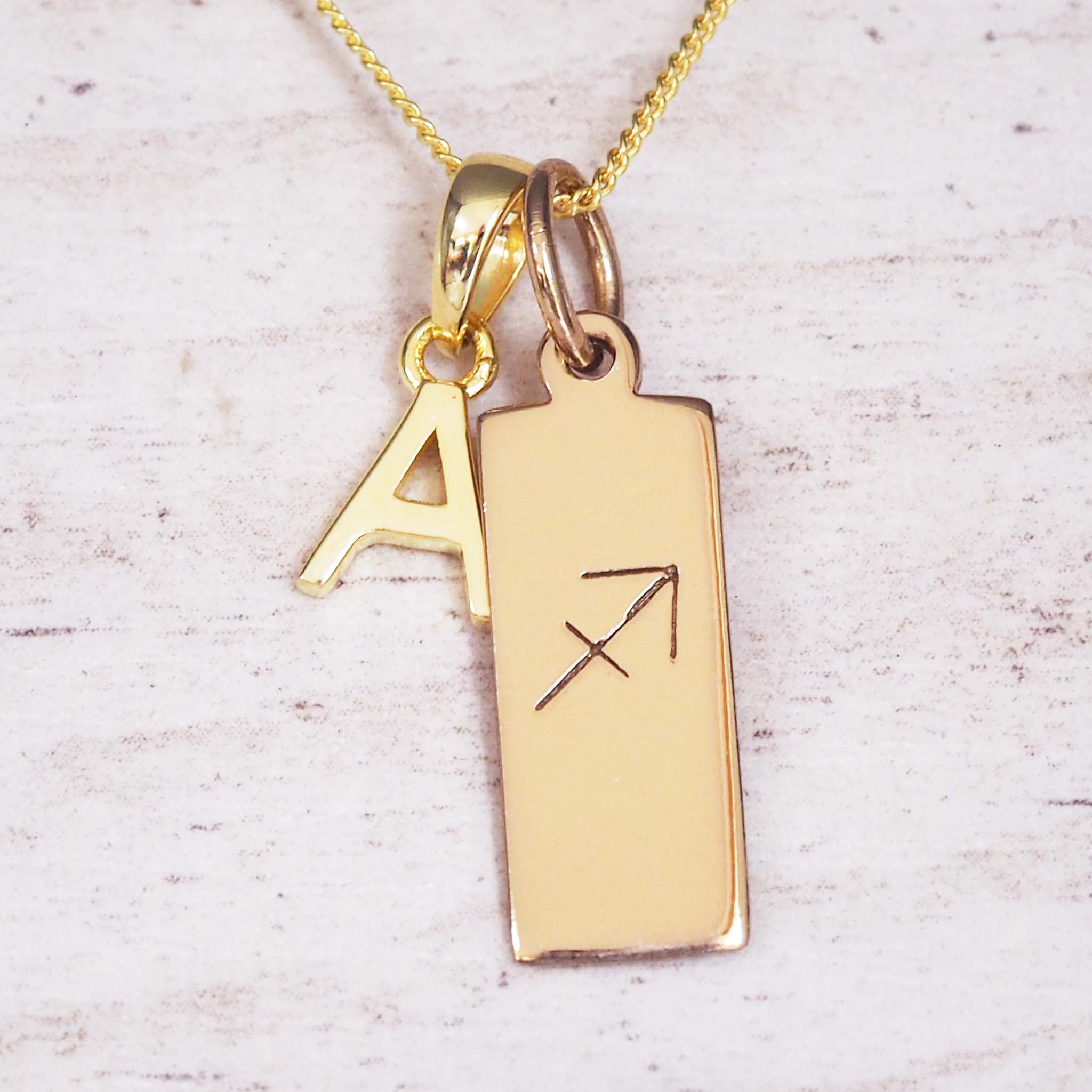 Gold necklace featuring an uppercase 'A' charm and a rectangular pendant engraved with the Sagittarius arrow glyph.