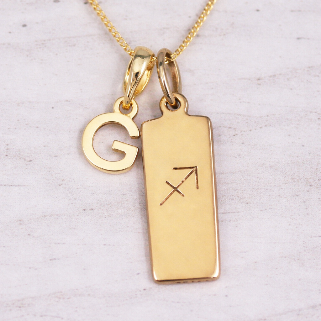 Gold necklace with 'G' pendant and rectangular tag with astrological symbol on a light background