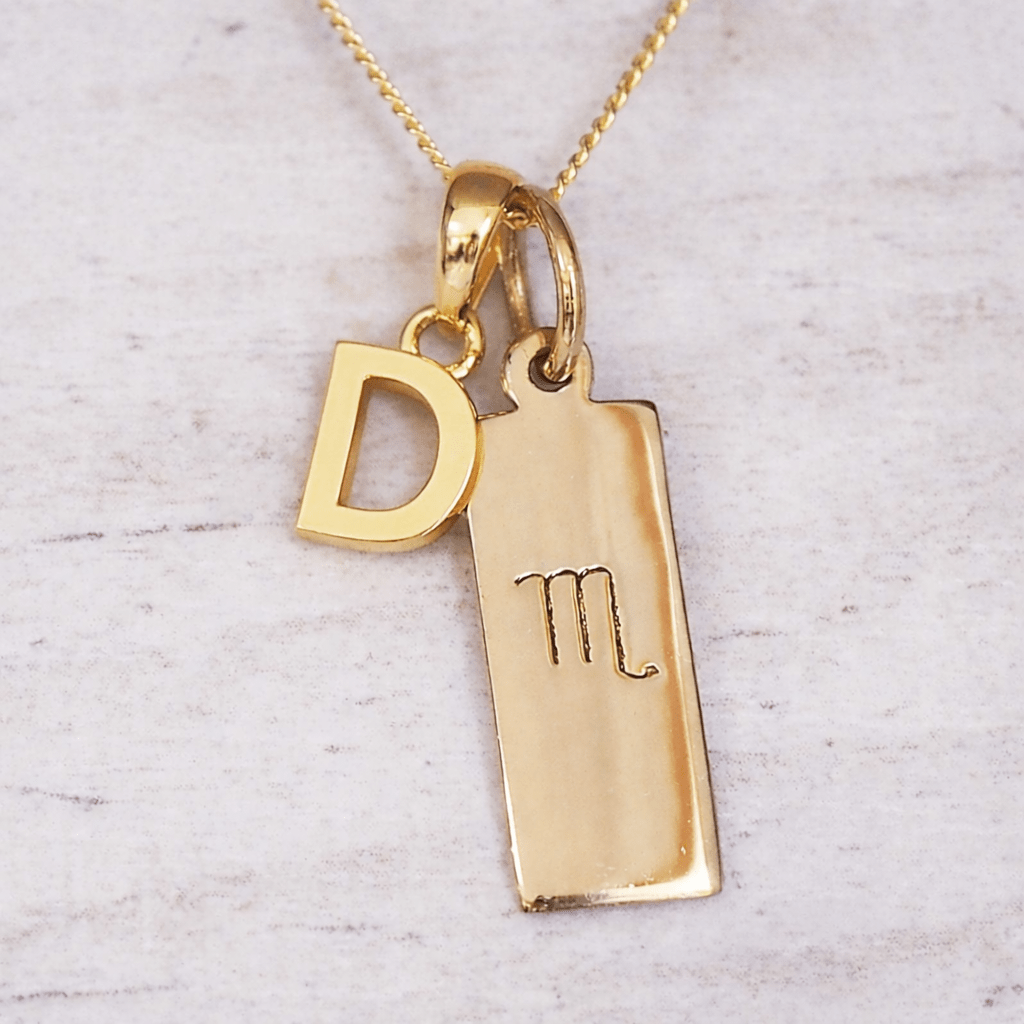 Gold necklace with 'D' initial pendant and scorpio zodiac pendant on a light gray background