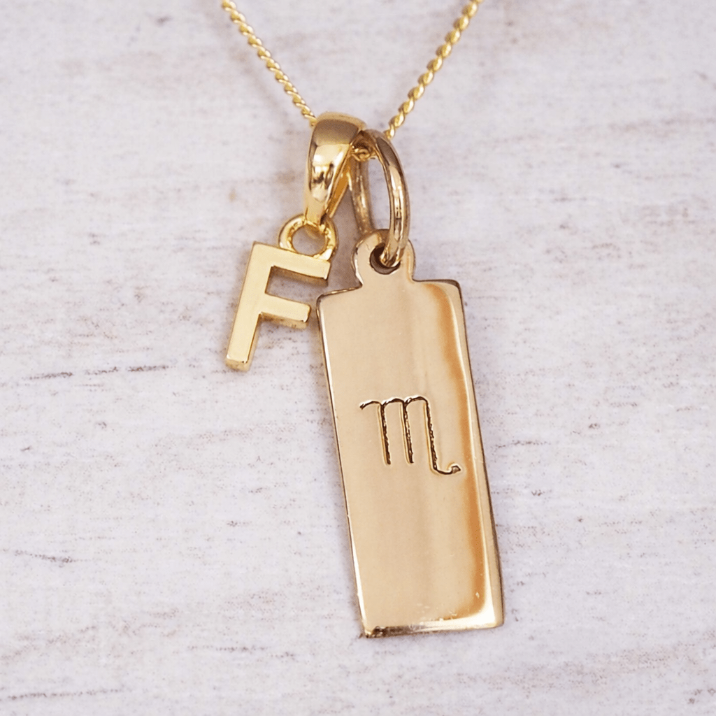 Gold necklace with a rectangular scorpio zodiac pendant and 'F' initial charm on a light background