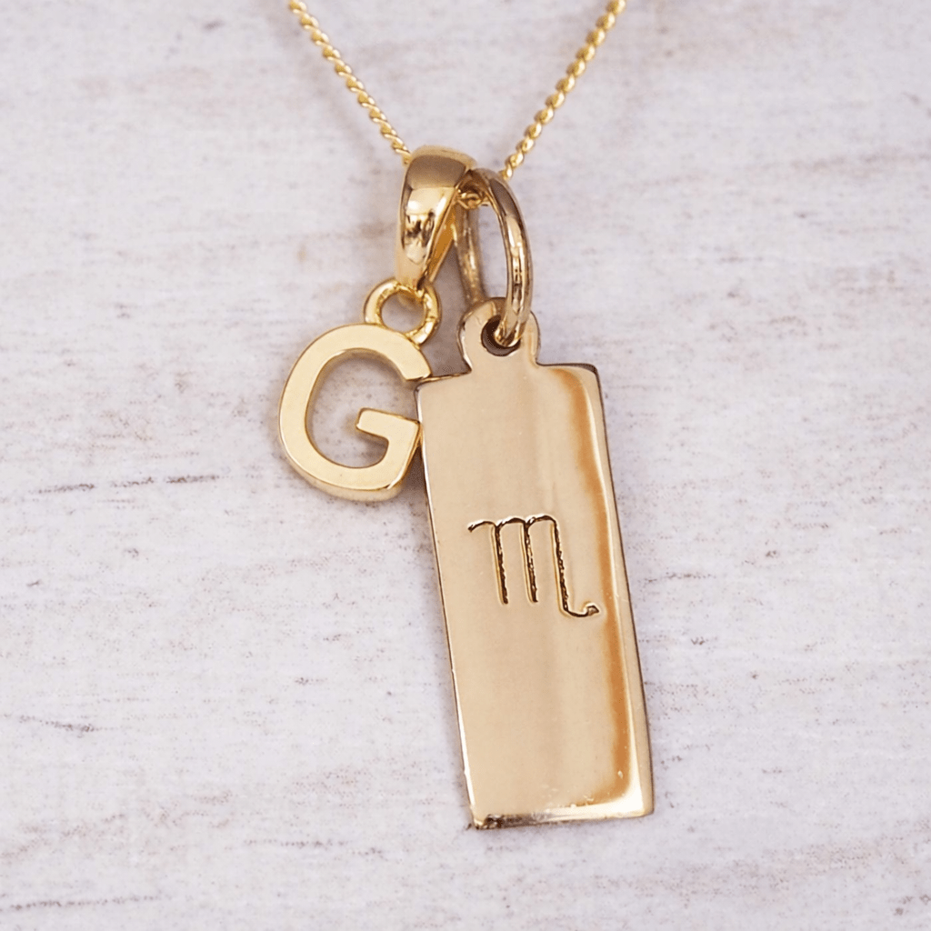 Gold necklace with a pendant featuring the letter 'G' and an astrological scorpio symbol on a light gray background.