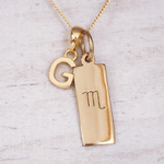 Gold necklace with a pendant featuring the letter 'G' and an astrological scorpio symbol on a light gray background.