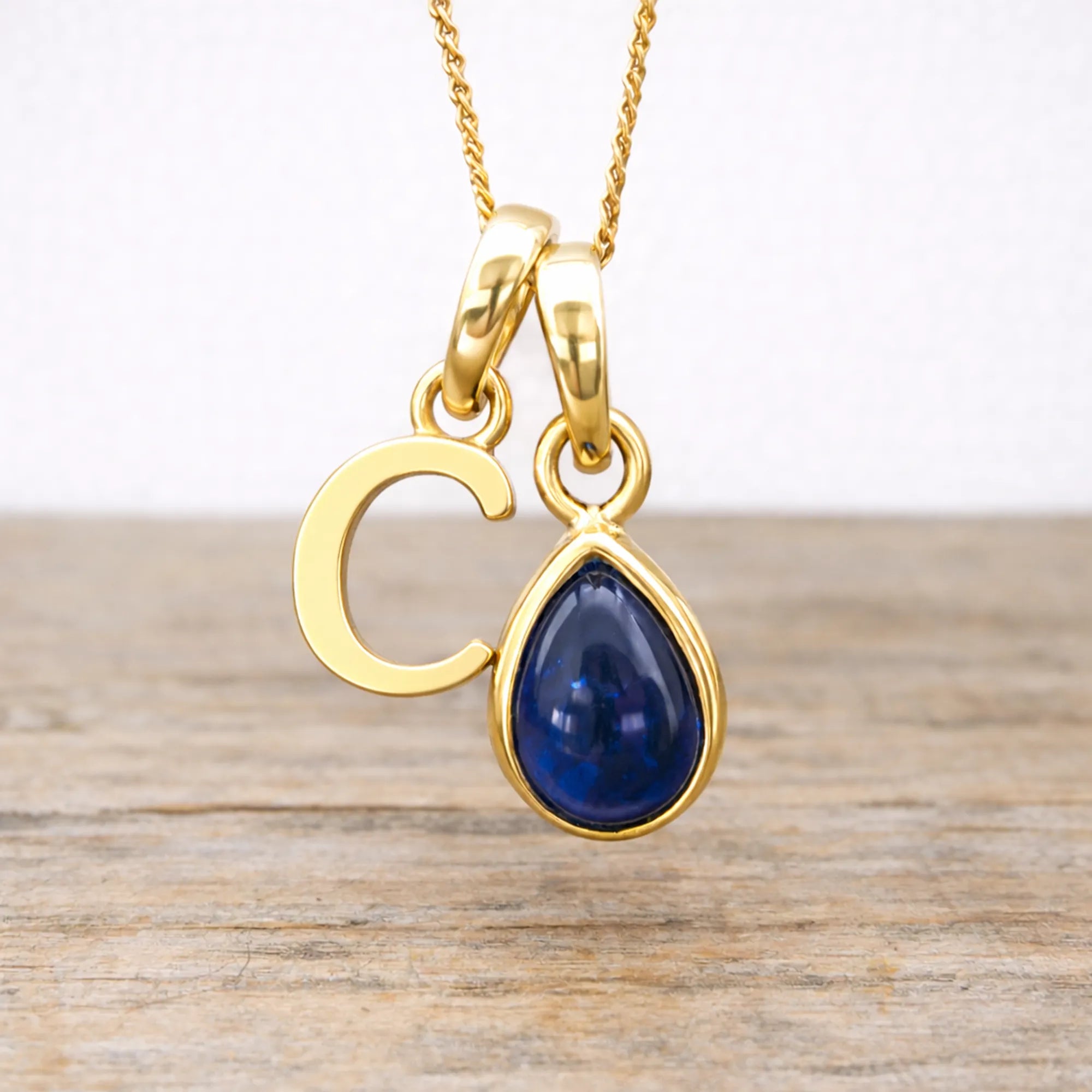 Gold September birthstone necklace with a blue sapphire teardrop pendant and letter 'C' on a wooden surface