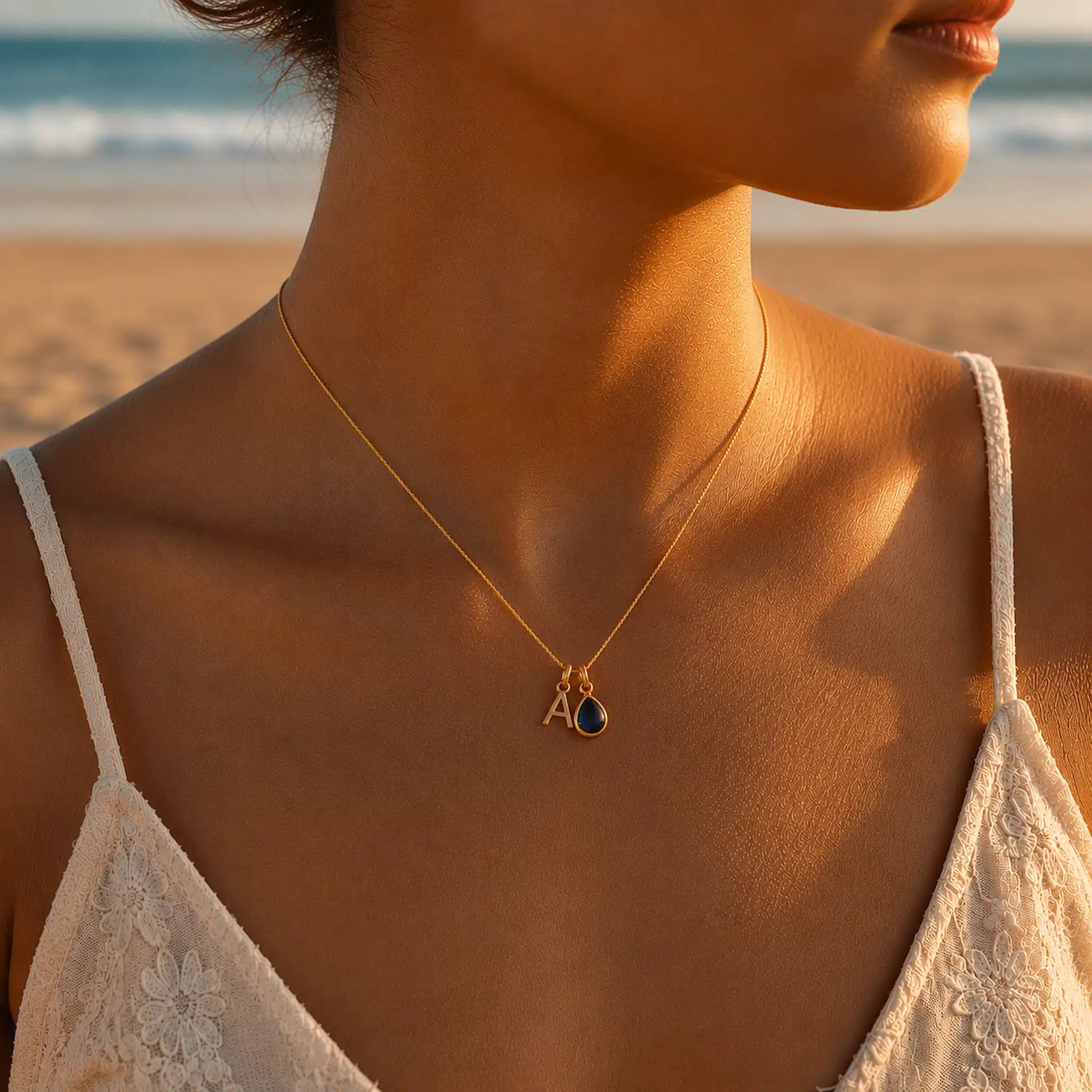 Woman wearing a fine gold chain necklace with a pear-shaped September Birthstone Sapphire pendant and a uppercase A letter pendant on a beach