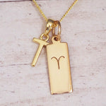 Gold initial T and Aries zodiac bar necklace, close-up of the letter charm and engraved pendant on whitewashed wood