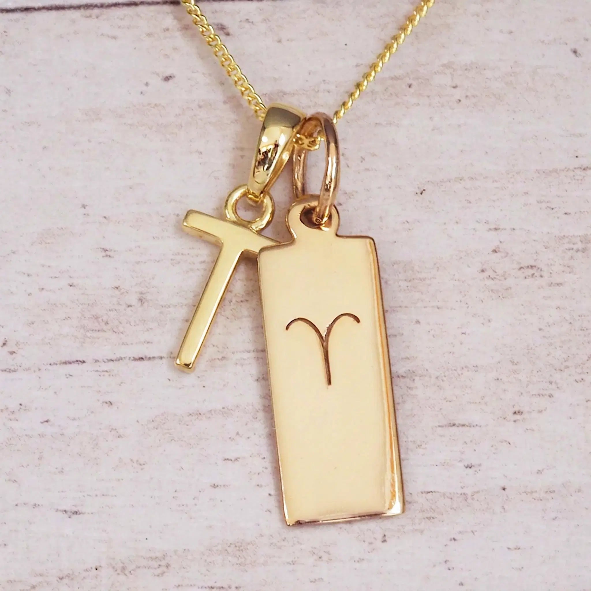 Gold initial T and Aries zodiac bar necklace, close-up of the letter charm and engraved pendant on whitewashed wood