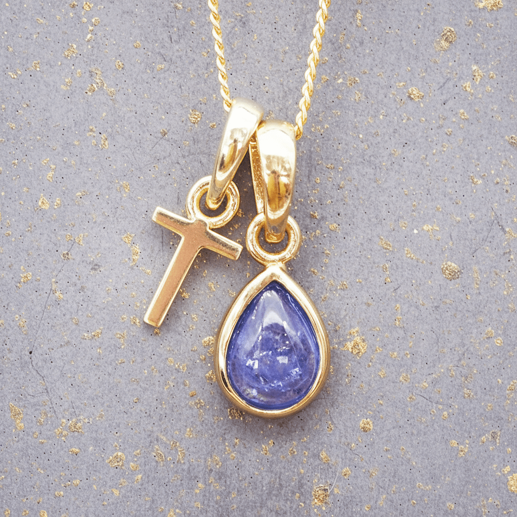 Gold necklace with a 'T' initial pendant and a teardrop-shaped purple december birthstone tanzanite gemstone on a gray background.