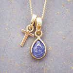 Gold necklace with a 'T' initial pendant and a teardrop-shaped purple december birthstone tanzanite gemstone on a gray background.