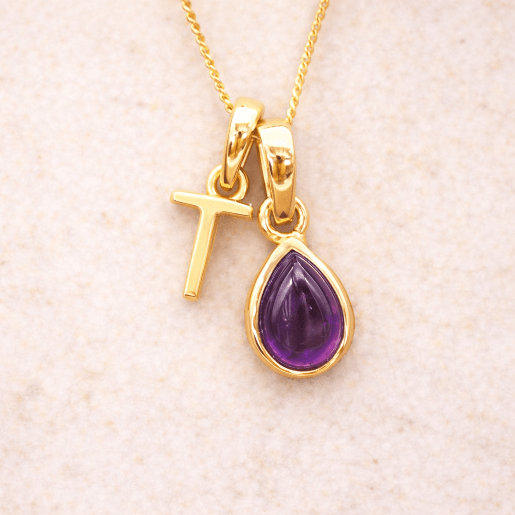 Gold necklace with a purple teardrop amethyst pendant and letter 'T' charm on a beige fabric background