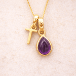 Gold necklace with a purple teardrop amethyst pendant and letter 'T' charm on a beige fabric background