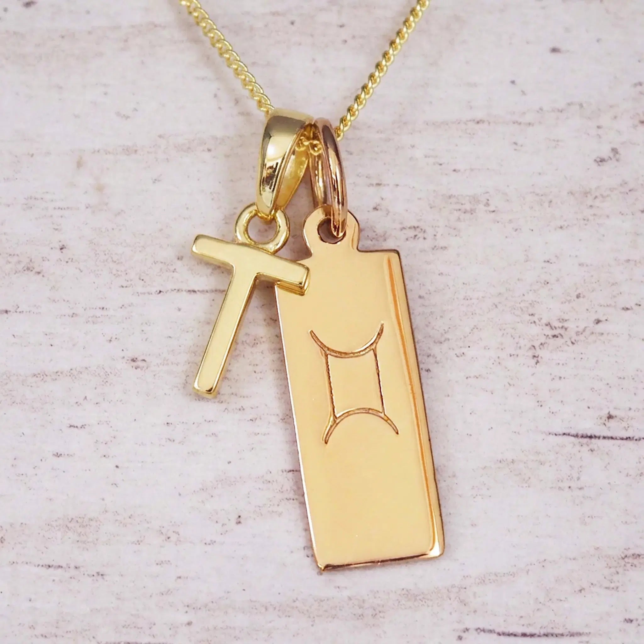 Gold initial T and Gemini zodiac bar necklace, close-up of the letter charm and engraved pendant on whitewashed wood