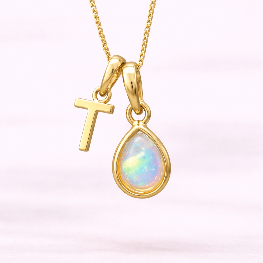 Gold necklace with a 'T' initial pendant and a teardrop-shaped opal pendant on a light pink background