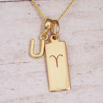 Gold initial U and Aries zodiac bar necklace, close-up of the letter charm and engraved pendant on whitewashed wood