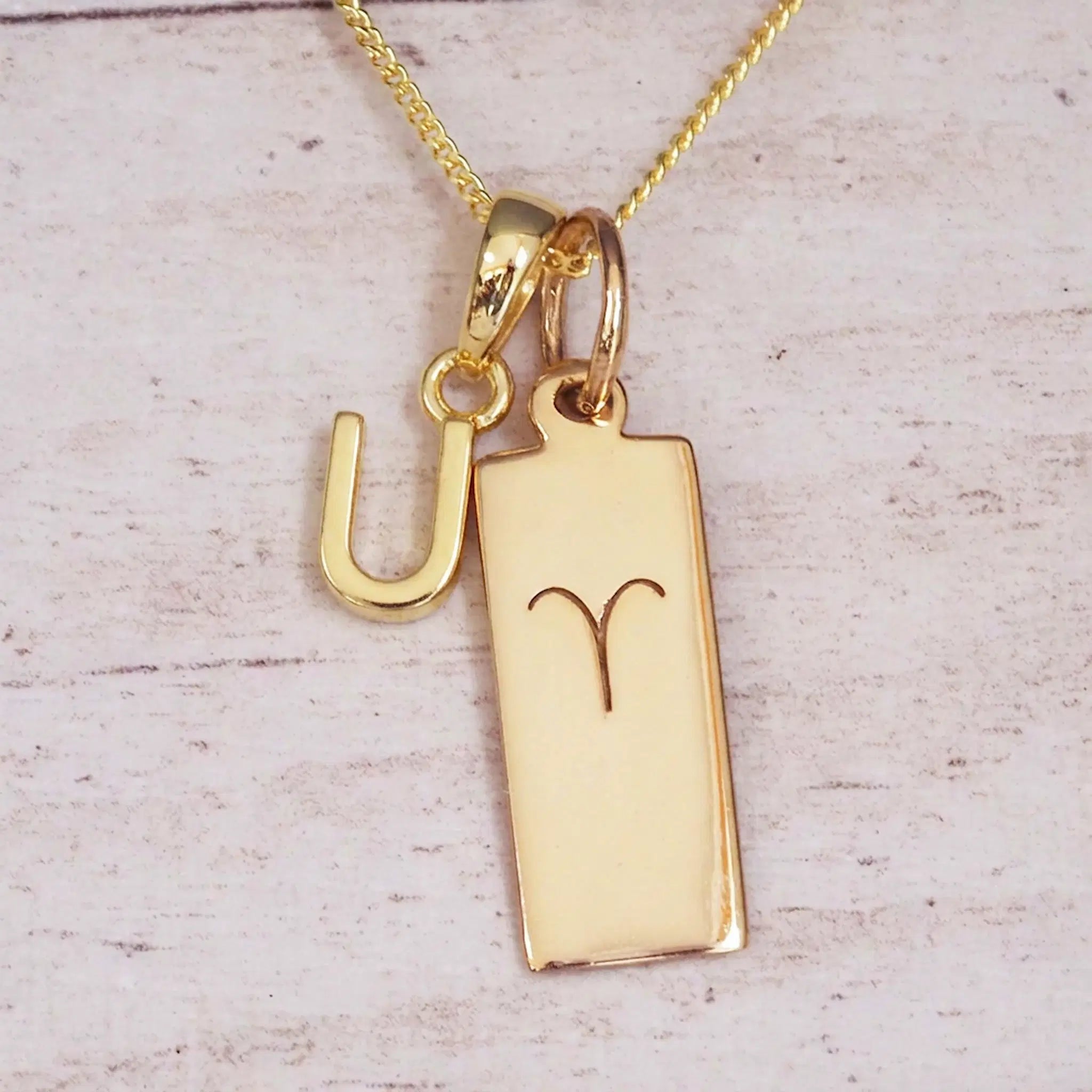 Gold initial U and Aries zodiac bar necklace, close-up of the letter charm and engraved pendant on whitewashed wood