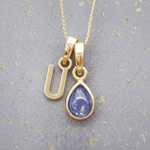 Gold necklace with a 'U' initial pendant and a purple teardrop december birthstone tanzanite pendant on a textured grey background