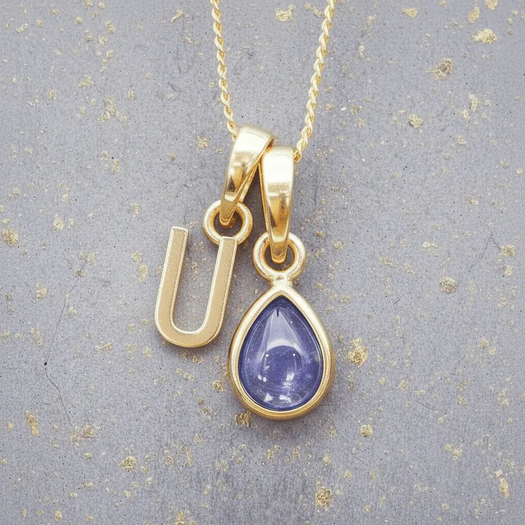 Gold December birthstone necklace with a pear-shaped natural tanzanite pendant and letter 'U' charm with a grey background.
