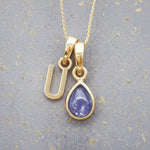 Gold December birthstone necklace with a pear-shaped natural tanzanite pendant and letter 'U' charm with a grey background.