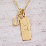 Gold initial U and Gemini zodiac bar necklace, close-up of the letter charm and engraved pendant on whitewashed wood