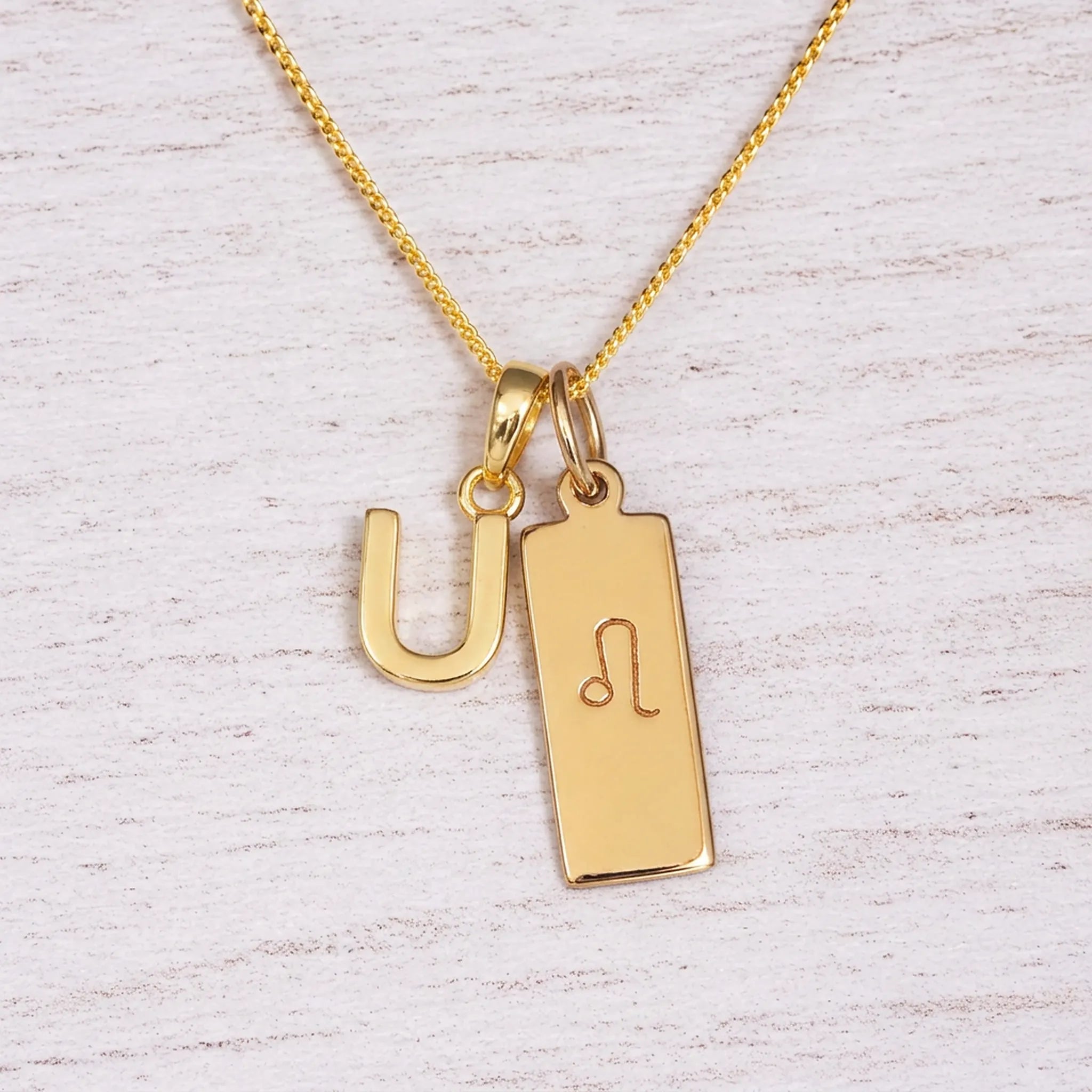 Gold initial and Leo zodiac necklace with a letter U pendant and engraved Leo bar on white wood, close-up