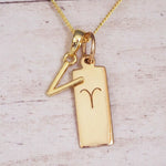 Gold initial V and Aries zodiac bar necklace, close-up of the letter charm and engraved pendant on whitewashed wood