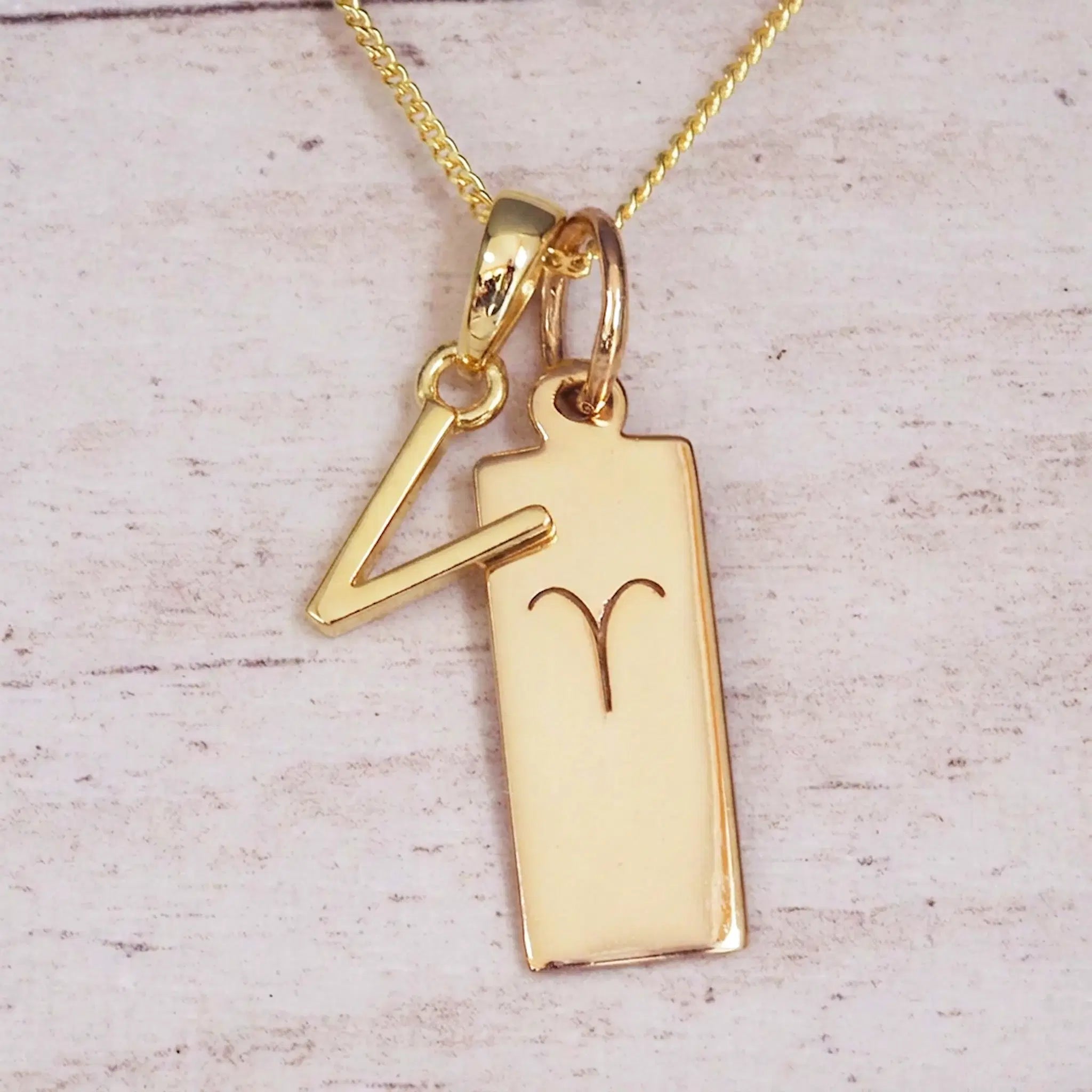 Gold initial V and Aries zodiac bar necklace, close-up of the letter charm and engraved pendant on whitewashed wood