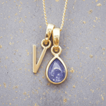 Gold necklace with 'V' initial pendant and teardrop-shaped purple december birthstone tanzanite gemstone on a grey background